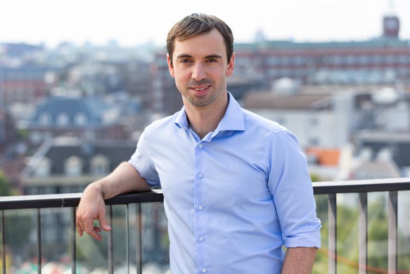 Booking.com chief business officer James Waters wears a light blue button-up shirt and leans on a rooftop railing with a blurred city backdrop.